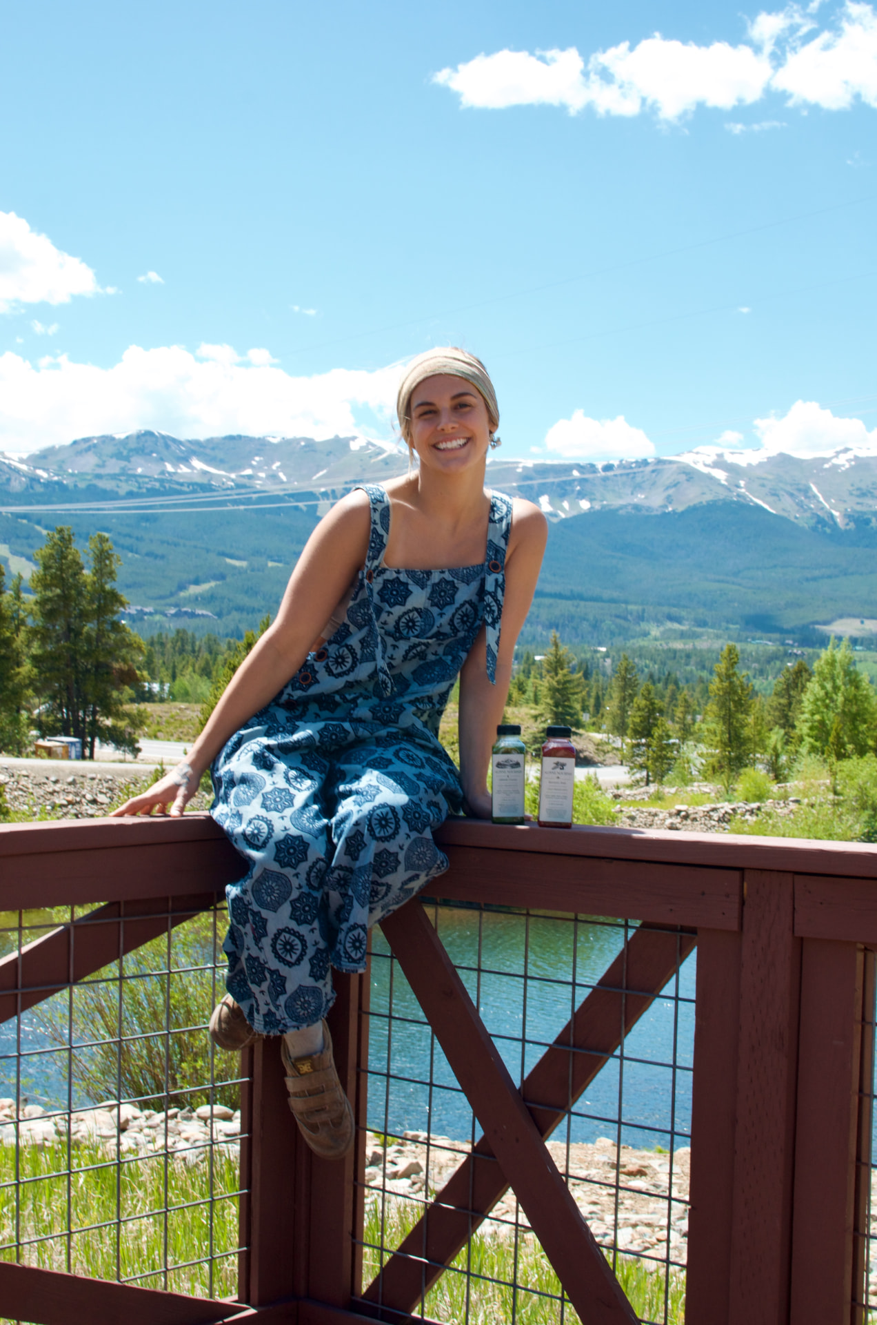 McKenzie, founder of Alpine Nourish, sitting on a wooden railing with snow-dusted mountains and pine trees behind her